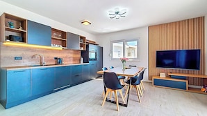 A modern kitchen with blue cabinets, a dining area with a wooden table and chairs, and a flat-screen TV mounted on a wooden panel wall.