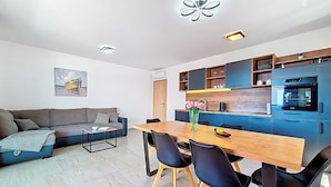 A modern living room with a sofa, coffee table, and dining area with chairs and a table. There is a kitchen with blue cabinets and a stove.
