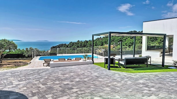 A modern outdoor pool area with a clear view of the sea, a covered seating area, and a well-maintained lawn.