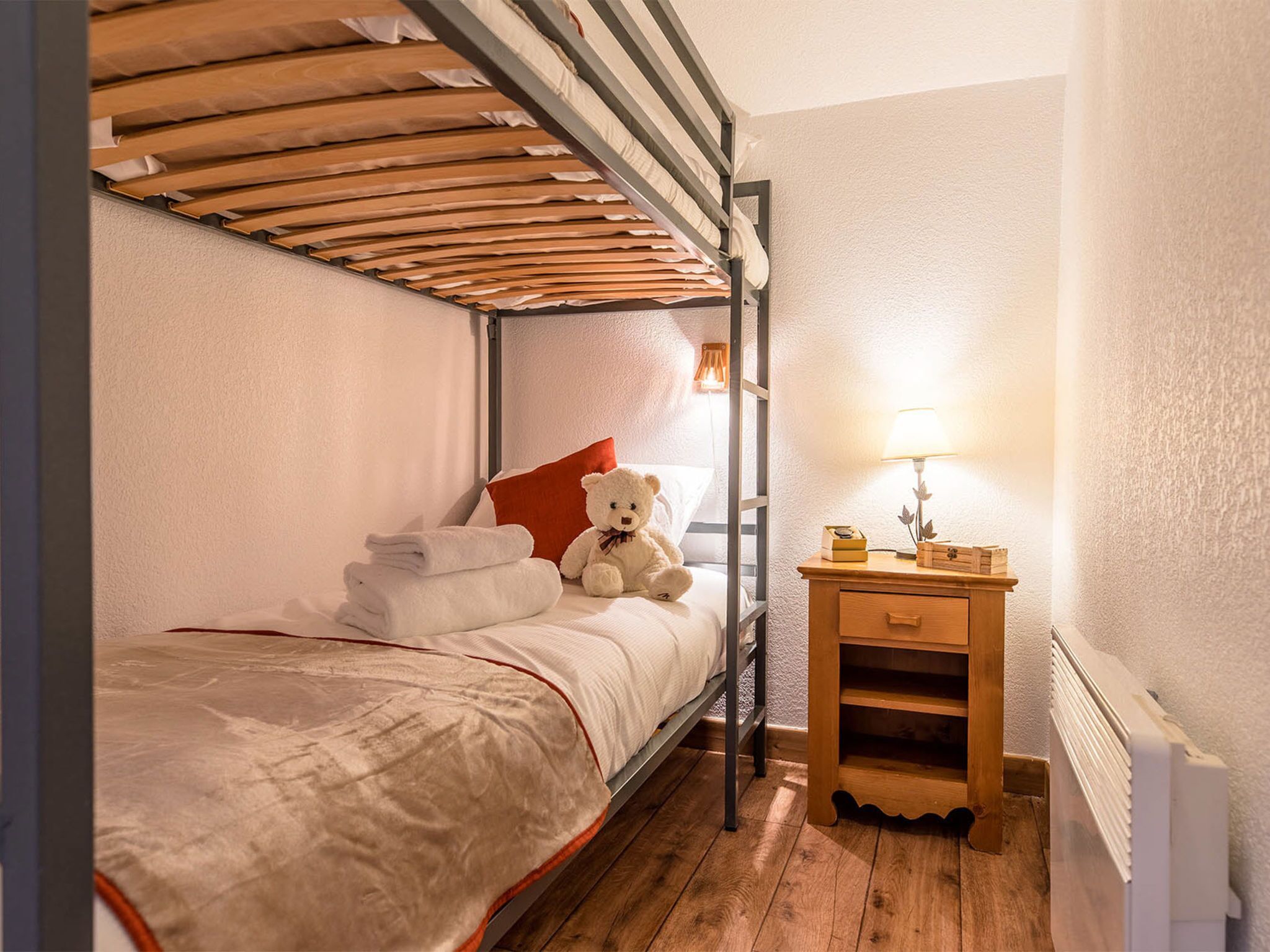 Una cama litera con cabecera de madera, una mesita de noche con lámpara y un osito de peluche sobre la cama.