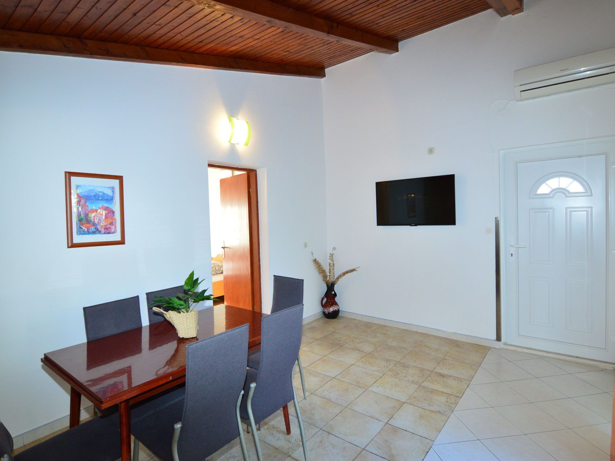Uma área de refeições com mesa e cadeiras de madeira, uma TV fixada na parede e uma porta com unidade de ar condicionado.