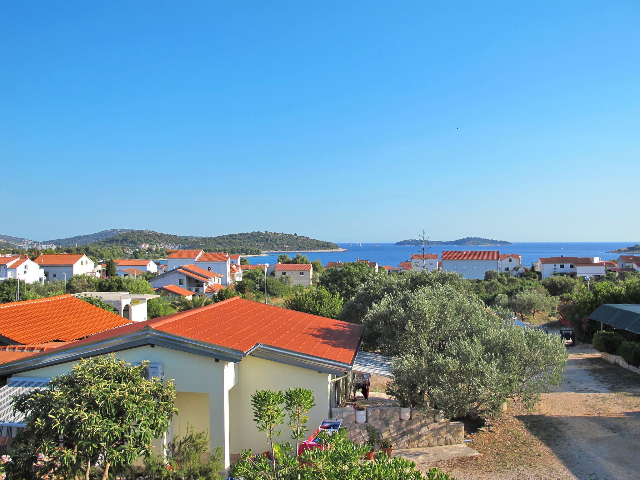 A coastal residential area with houses featuring red-tiled roofs, surrounded by greenery and a clear view of the sea.