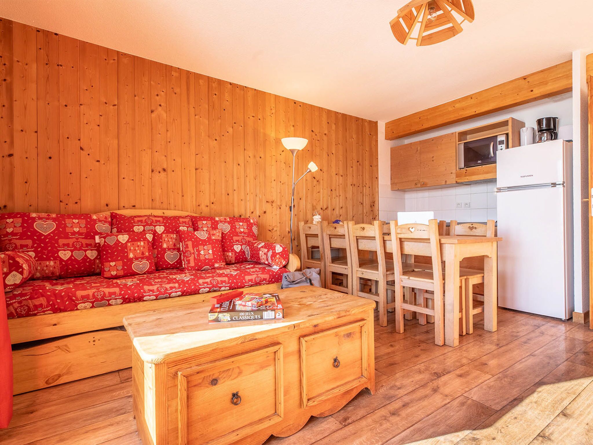 A cozy room with a wooden wall, a red patterned sofa, a wooden coffee table, and a kitchen area with a microwave and a refrigerator.