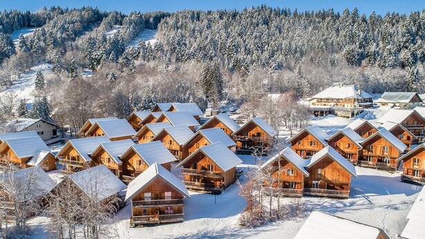 Eine Gruppe Holzhütten mit schneebedeckten Dächern, umgeben von einem bewaldeten Gebiet und einem Skipisten im Hintergrund.