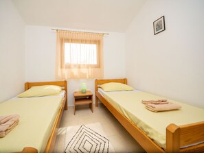 A twin room with two single beds, a small bedside table with a lamp, a window with sheer curtains, and a framed picture on the wall.