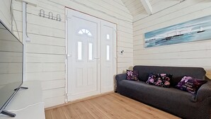 Una habitación con una puerta blanca, un sofá gris con cojines estampados, un cuadro de un barco en la pared y un suelo de madera.