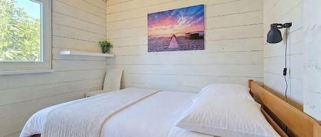 A bedroom with a wooden bed, a window, a wall-mounted shelf with a plant, and a sunset painting.