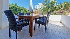 Un espacio al aire libre para comer con una mesa de madera y cuatro sillas de mimbre, una sombrilla blanca y vistas a edificios y árboles.