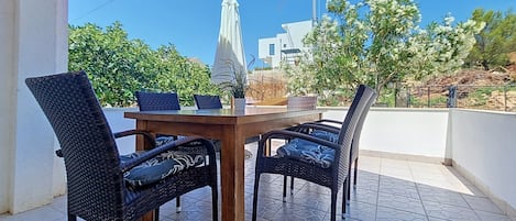 An outdoor dining area with a wooden table and four wicker chairs, a white umbrella, and a view of buildings and trees.