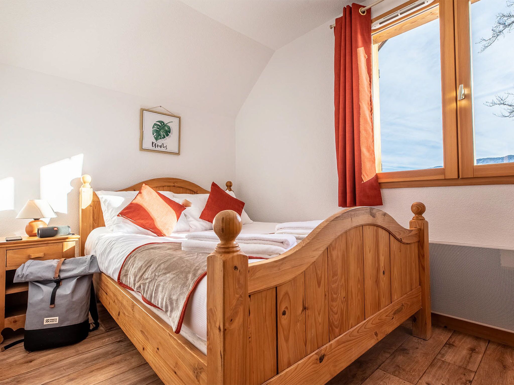 A wooden bed with red and white bedding, a bedside table with a lamp, a framed picture on the wall, and a window with curtains.