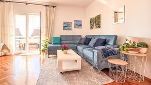 A modern living room with a grey sofa, a white coffee table, and a small round table with a plant. There is a sliding glass door leading to an outdoor area. - Apartment Ana by Interhome (Biograd na Moru)