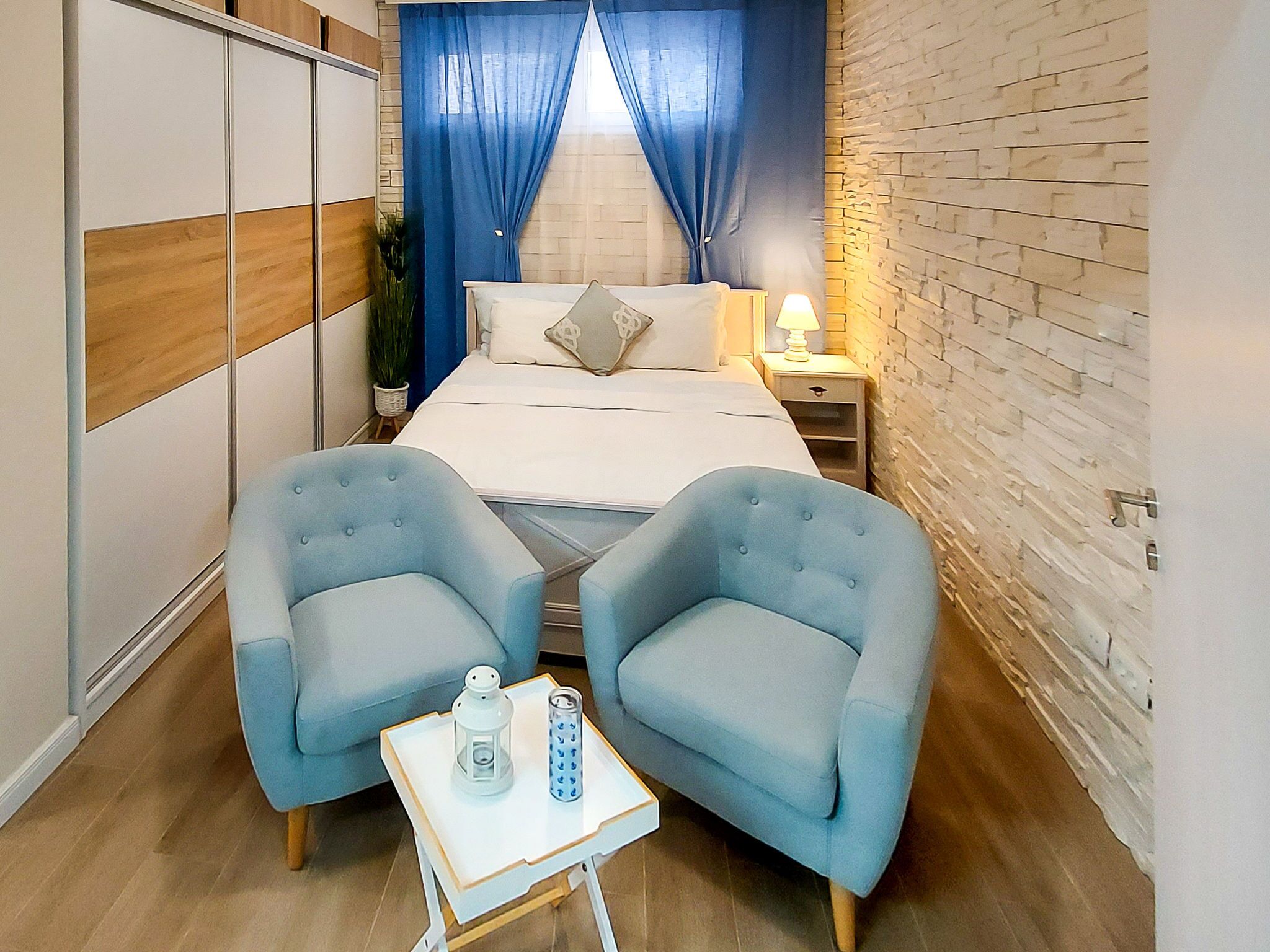 A bedroom with a bed, two blue armchairs, a small table with a bottle and a lantern, and a nightstand with a lamp.