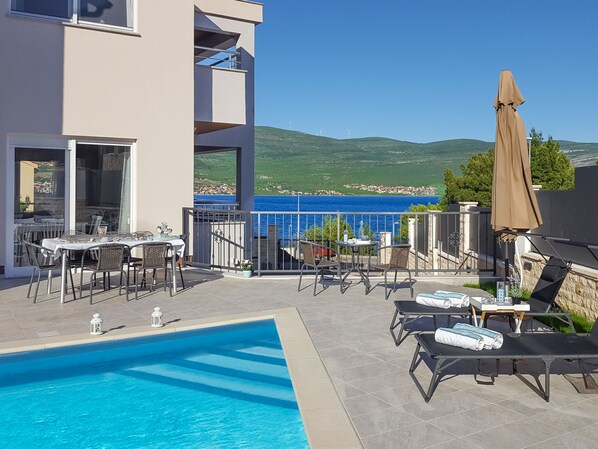 A poolside area with lounge chairs, a table, and a view of the sea and hills.