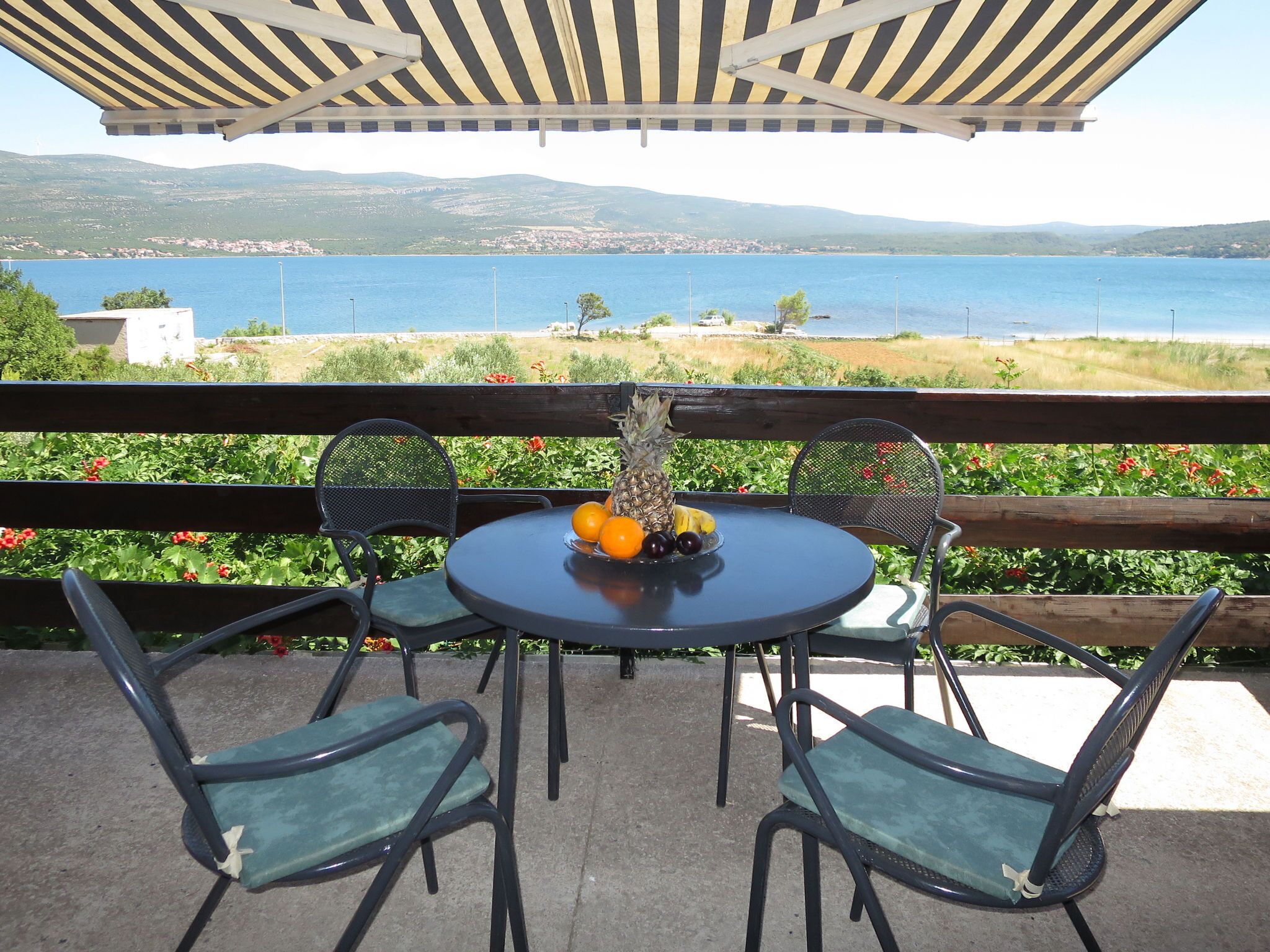 Ein Tisch mit Stühlen und einer Obstschale auf einem Balkon mit Blick auf ein Gewässer und Hügel.