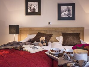 A neatly made bed with a red blanket, two pillows, a bedside lamp, a framed photograph, and a tray with bowls and a candle.
