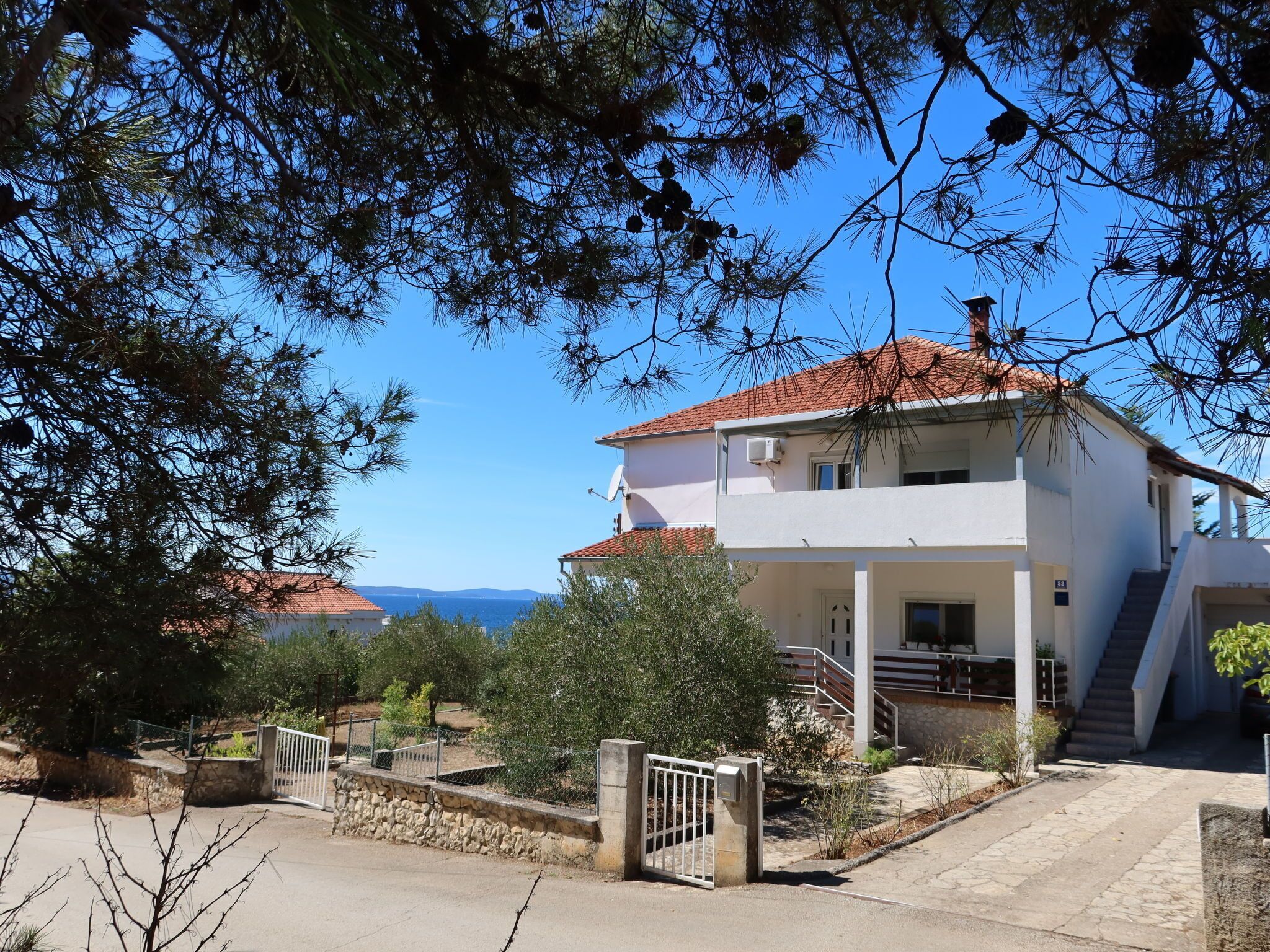 A two-story house with a red-tiled roof, a gated entrance, and a view of the sea in the background.