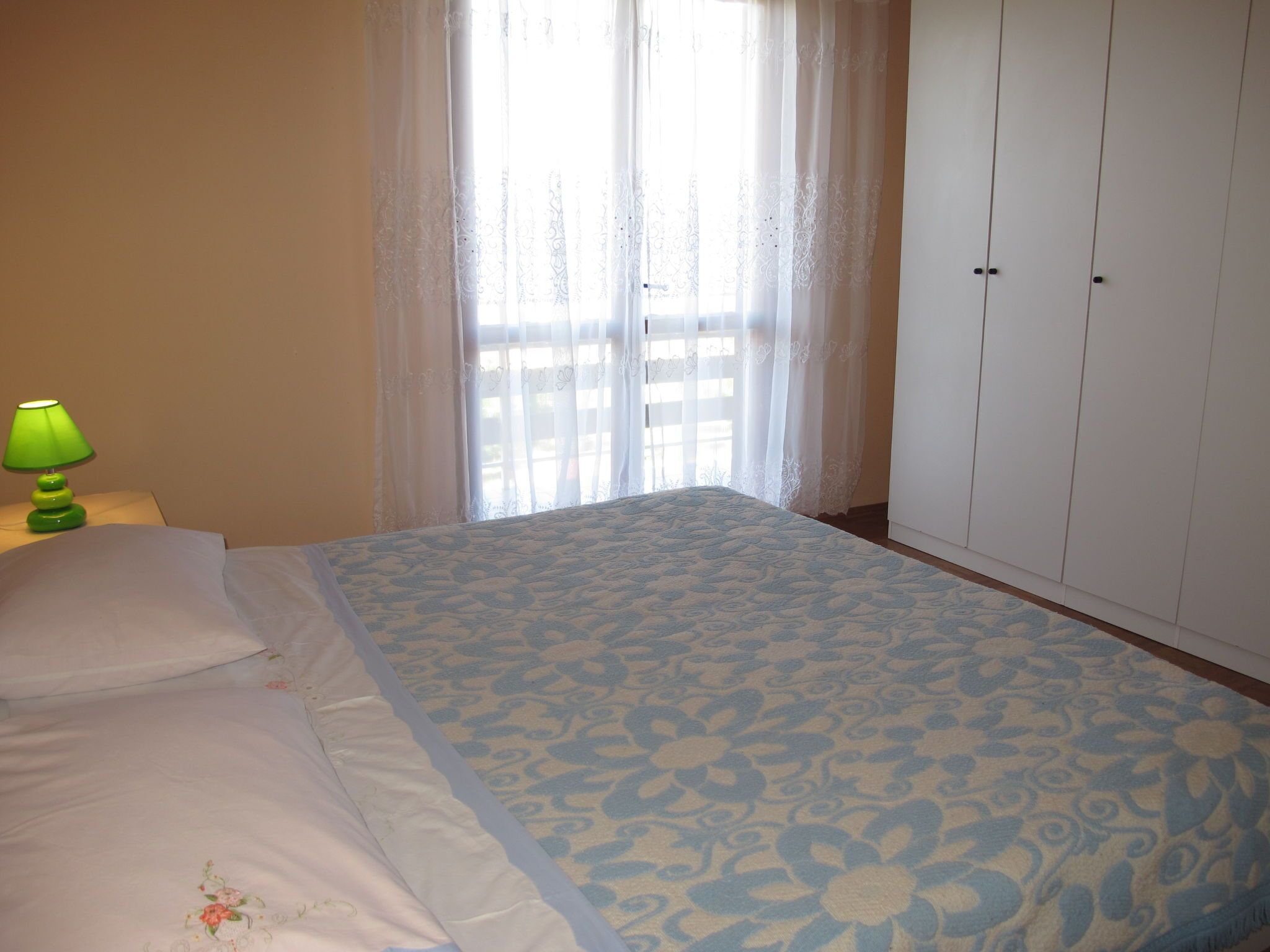 Un dormitorio con una cama, una lámpara verde, una ventana con cortinas traslúcidas y un armario.