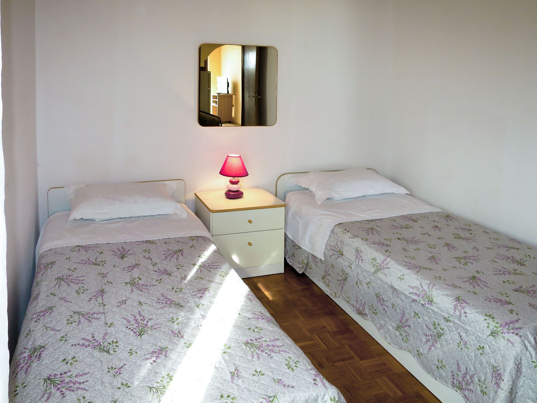 Een slaapkamer met twee enkele bedden, een nachtkastje met een lamp, een spiegel en een houten vloer.