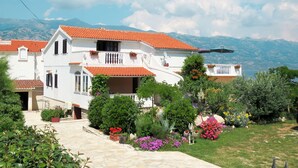 A two-story white building with red-tiled roofs, a stone pathway leading to the entrance, and a garden with various plants and flowers. - Apartment Lukeni by Interhome (Vinjerac)