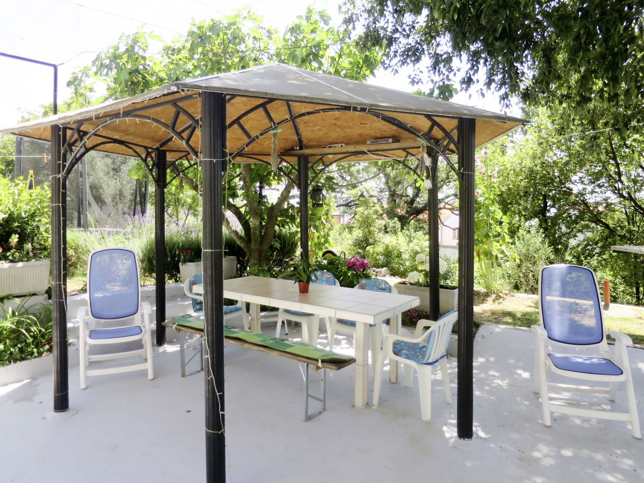 A covered outdoor seating area with a table and chairs, surrounded by greenery.