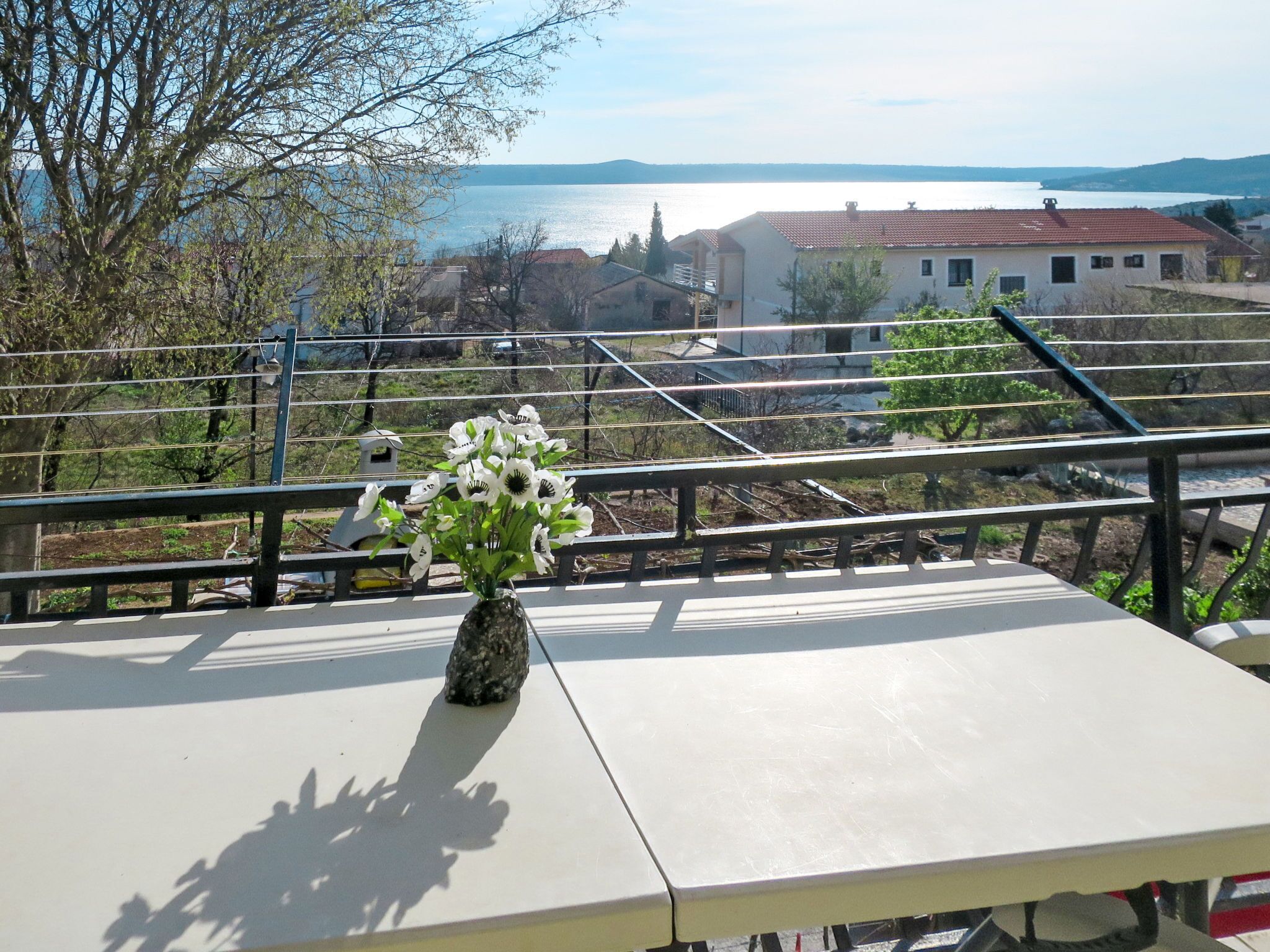 A balcony with a table and a vase of flowers, overlooking a body of water and buildings.