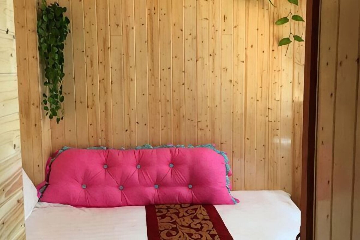 A room with a wooden paneled wall, a bed with a pink tufted headboard, and a green plant on the left.