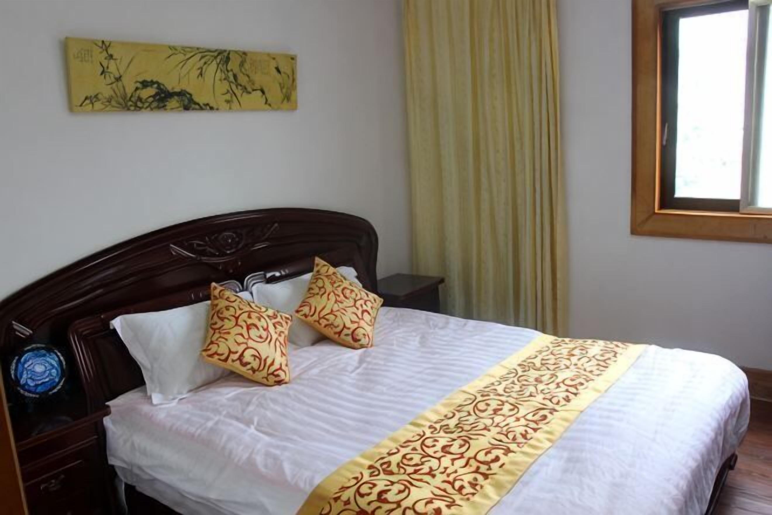 A neatly made bed with a patterned bedspread and two decorative pillows, a bedside table with a blue object, and a window with a yellow curtain.