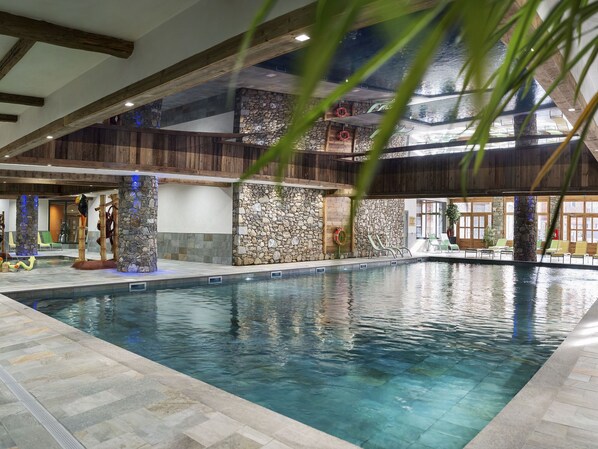 An indoor swimming pool with stone walls and wooden beams, surrounded by lounge chairs and a view of the sky.