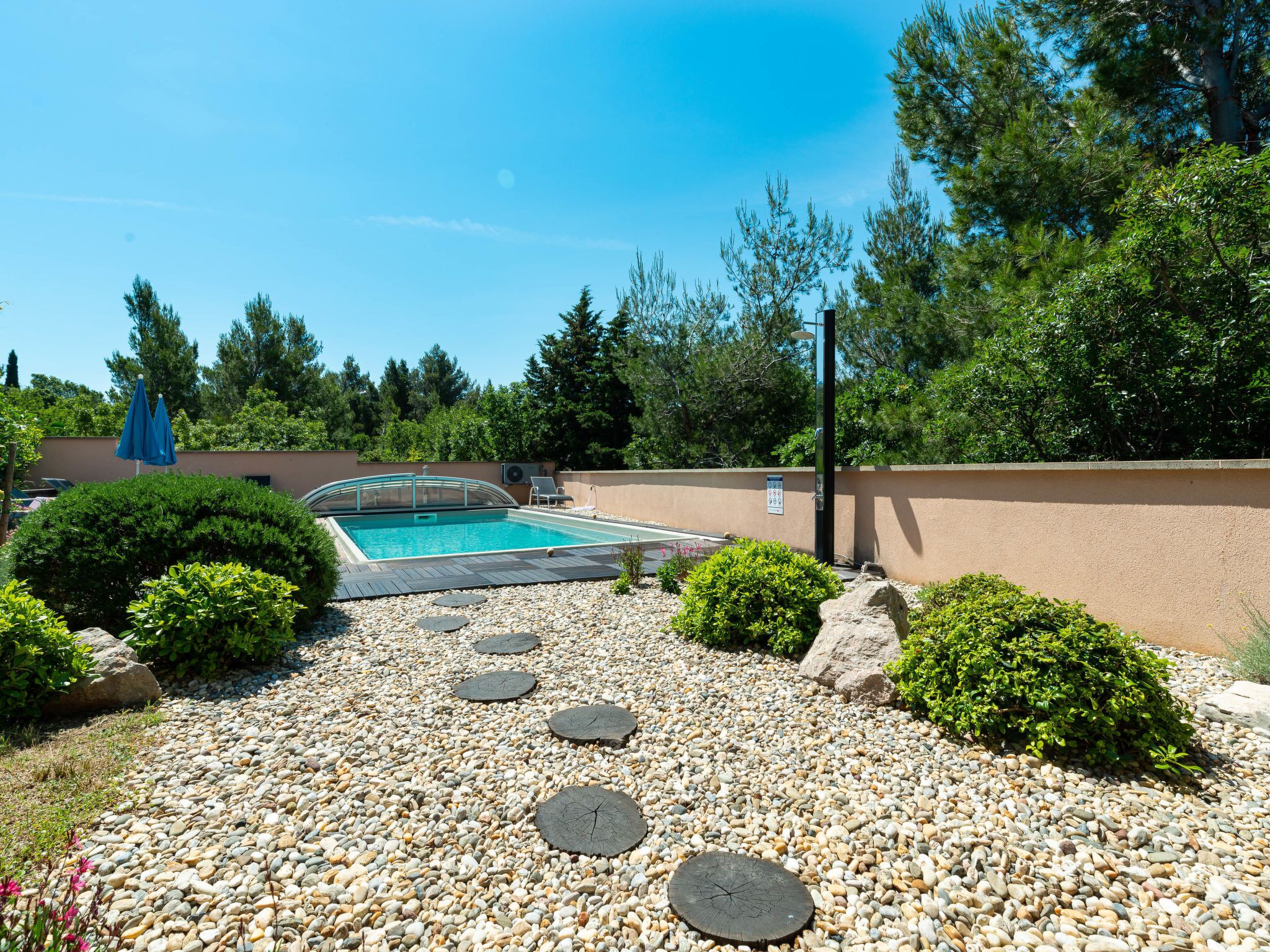A swimming pool with a pebble pathway, surrounded by greenery and a clear blue sky.