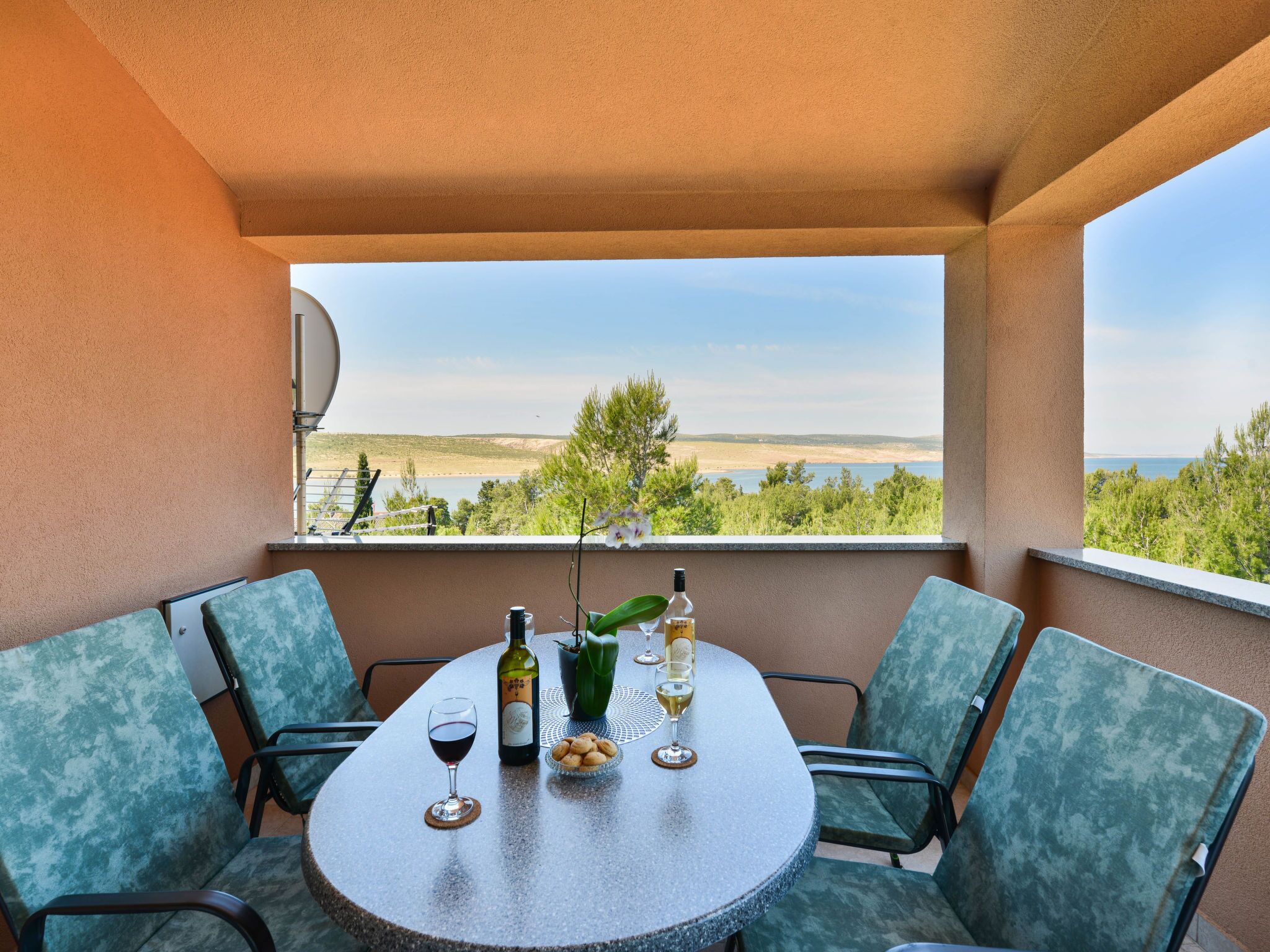 En balkong med ett runt bord dukat för två personer, med en vinflaska, glas och en liten skål med snacks. Från balkongen har man utsikt över ett vackert landskap med träd och en sjö eller ett annat vattendrag.