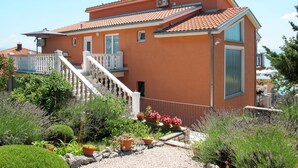 Uma casa de dois andares com telhado de telhas vermelhas, uma varanda com balaústres brancos e um jardim com diversas plantas e flores.