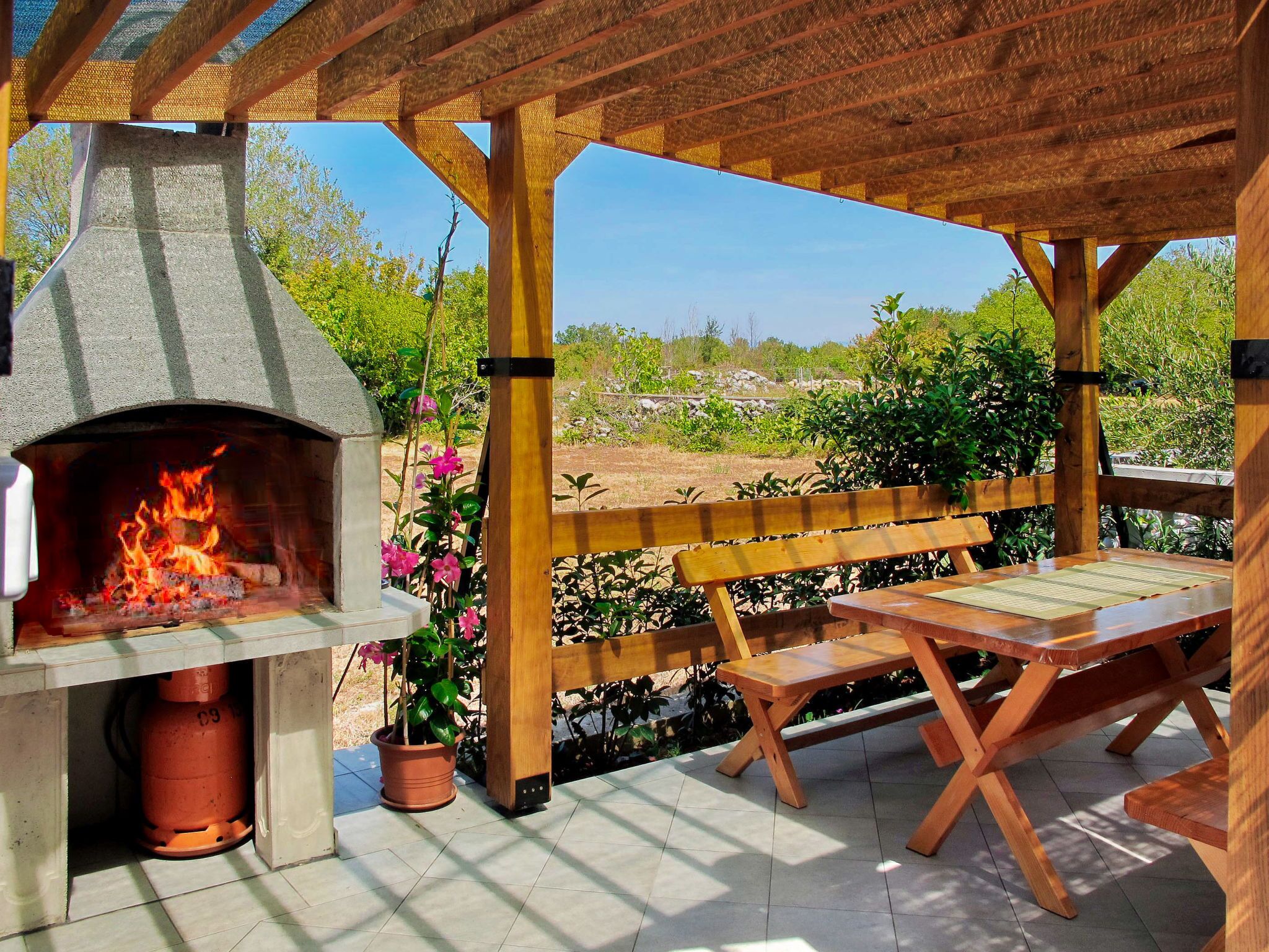 An outdoor patio with a stone-built wood-fired pizza oven, wooden benches, and a view of a natural landscape.