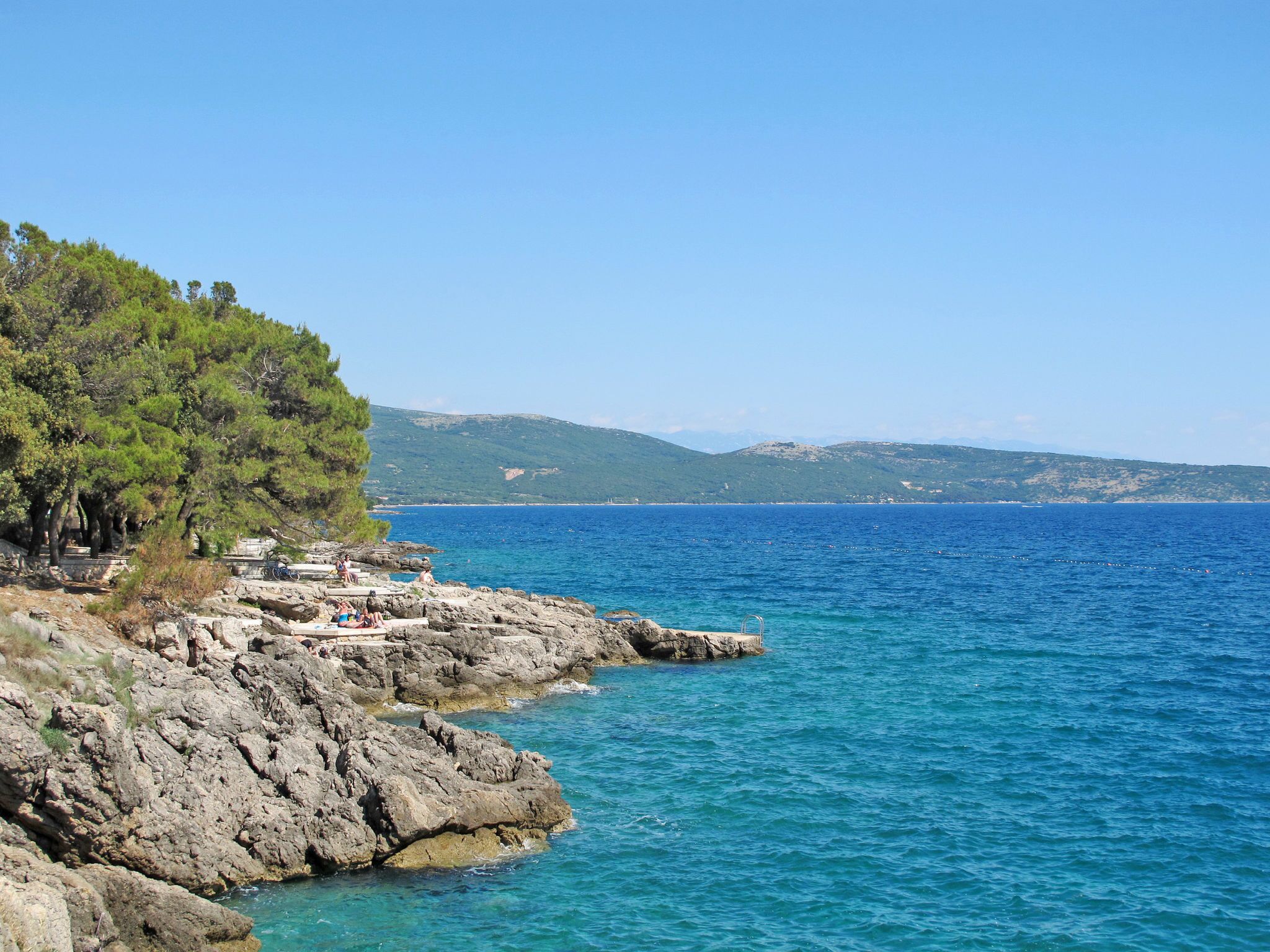 A rocky coastline with a clear blue sea, a few people, and a distant mountain range.