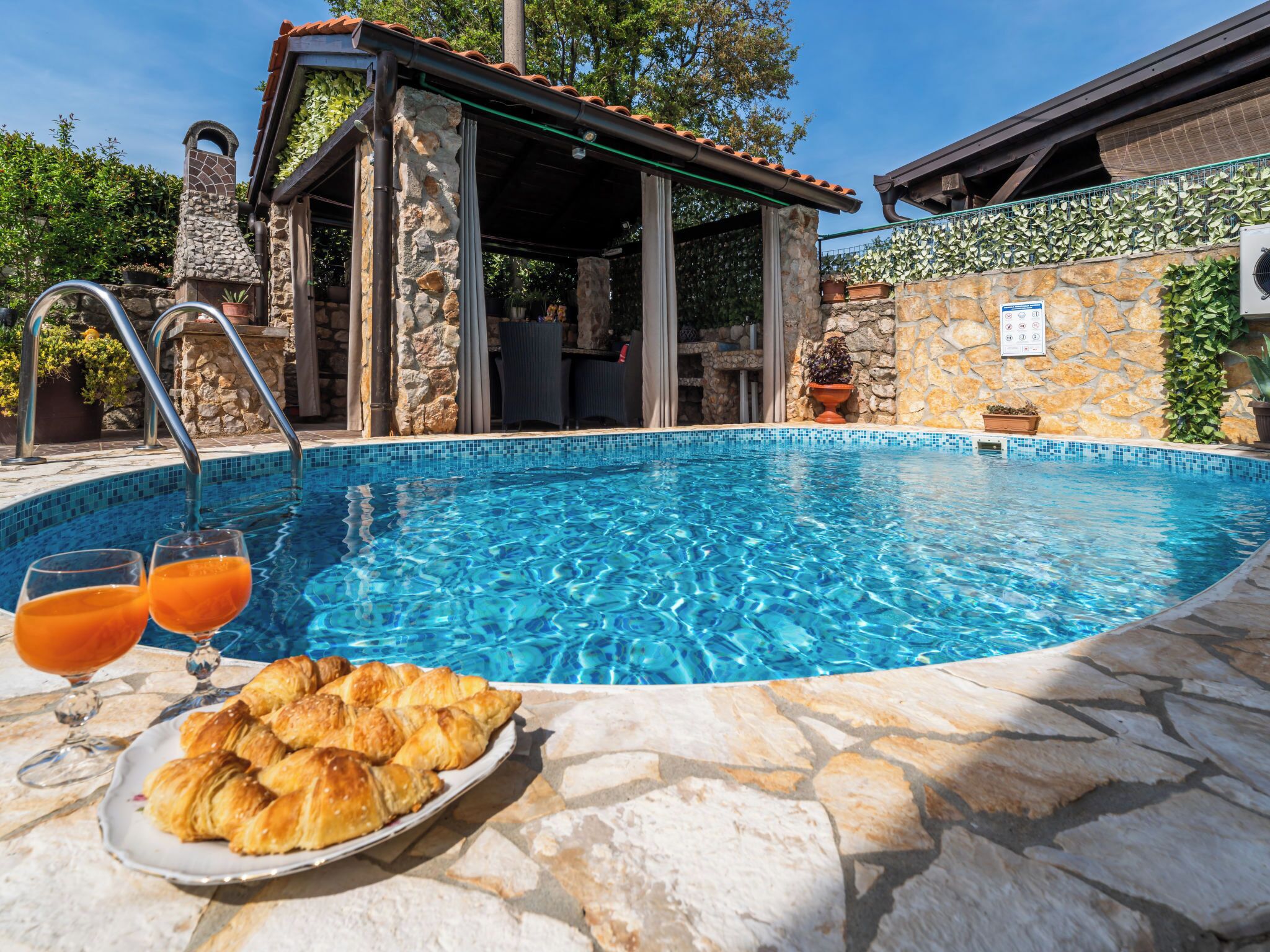 En pool med stenplattor, en möblerad uteplats under tak och en tallrik med croissanter.