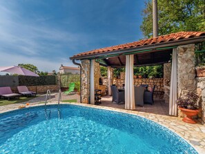 A swimming pool with a stone edge, outdoor seating, and a covered area with stone pillars.