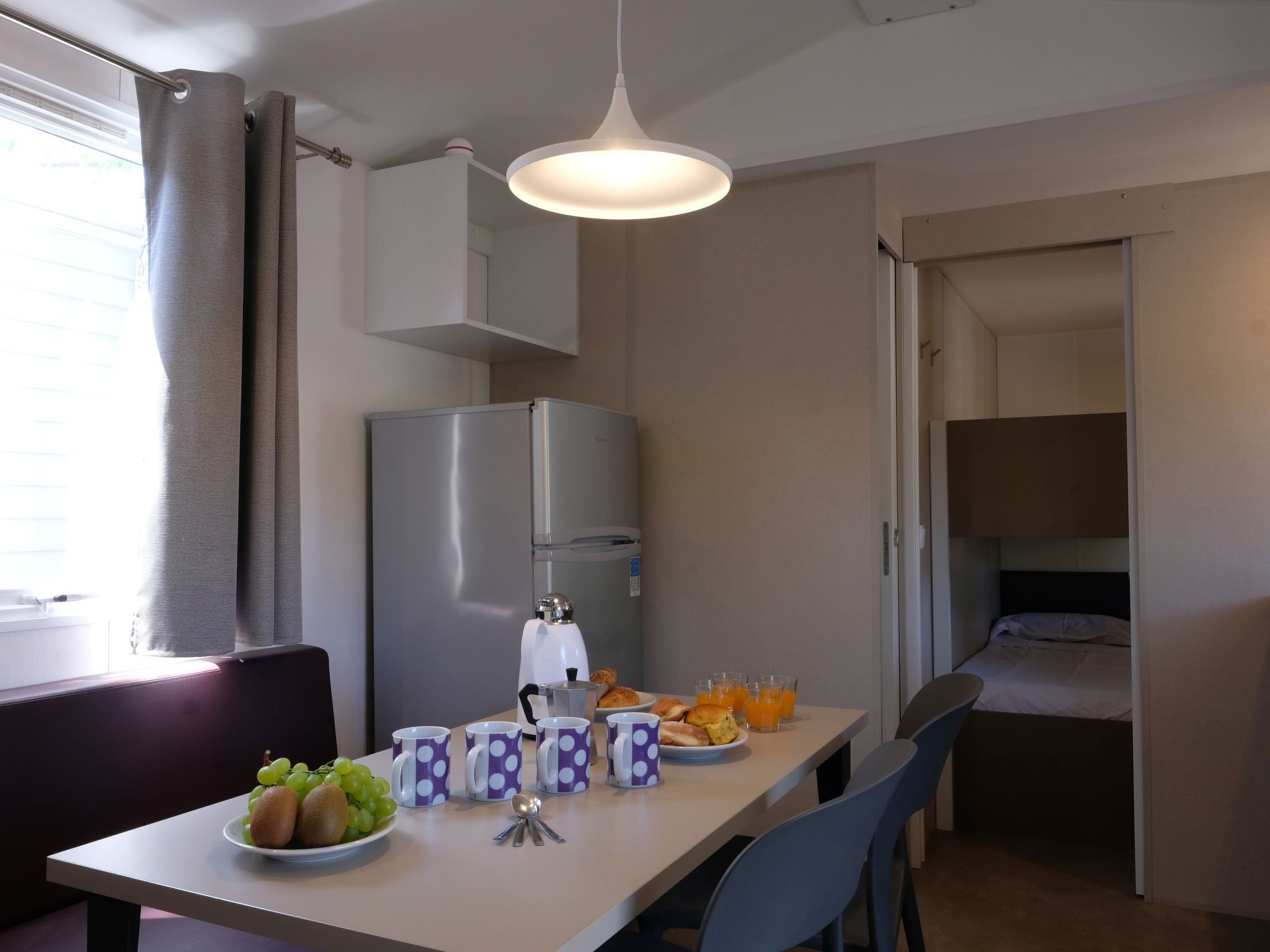 Cocina compacta con refrigerador, una mesa de comedor con productos para el desayuno y una cama litera al fondo.