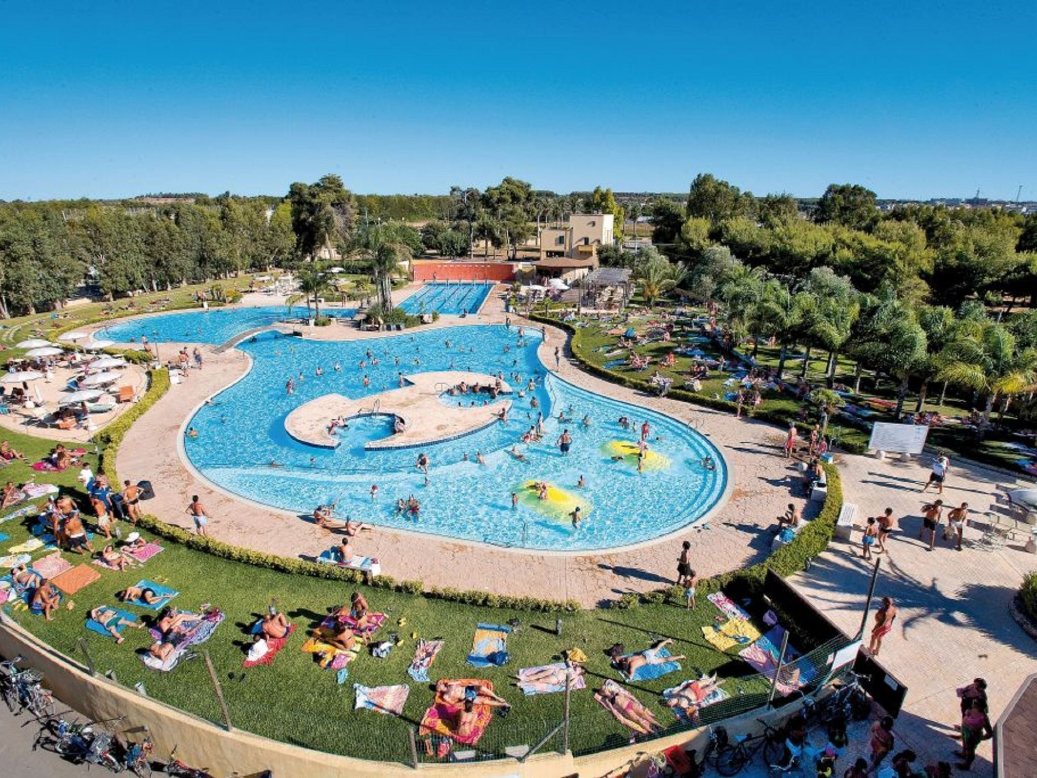 A large outdoor swimming pool with multiple lanes, surrounded by sun loungers and umbrellas, set in a landscaped area with trees and buildings in the background.