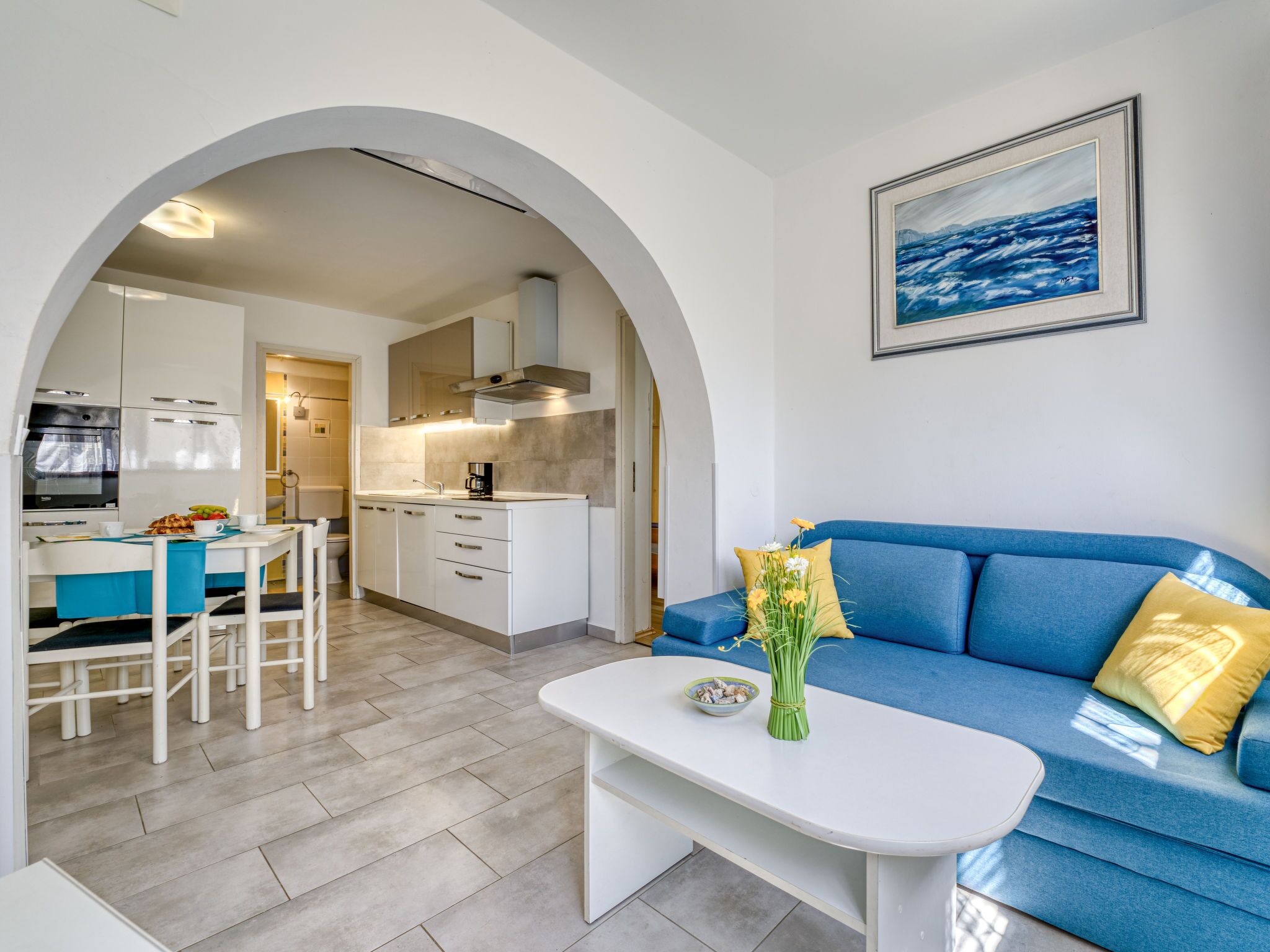 Eine moderne Küche mit weißen Schränken, ein Essbereich mit weißem Tisch und Stühlen sowie ein blaues Sofa mit gelben Kissen.