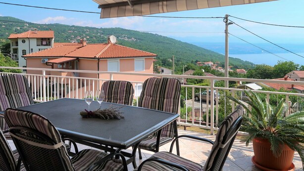 A balcony with a table and chairs, a potted plant, and a view of a hillside with houses. - Katarina by Interhome (Lovran)