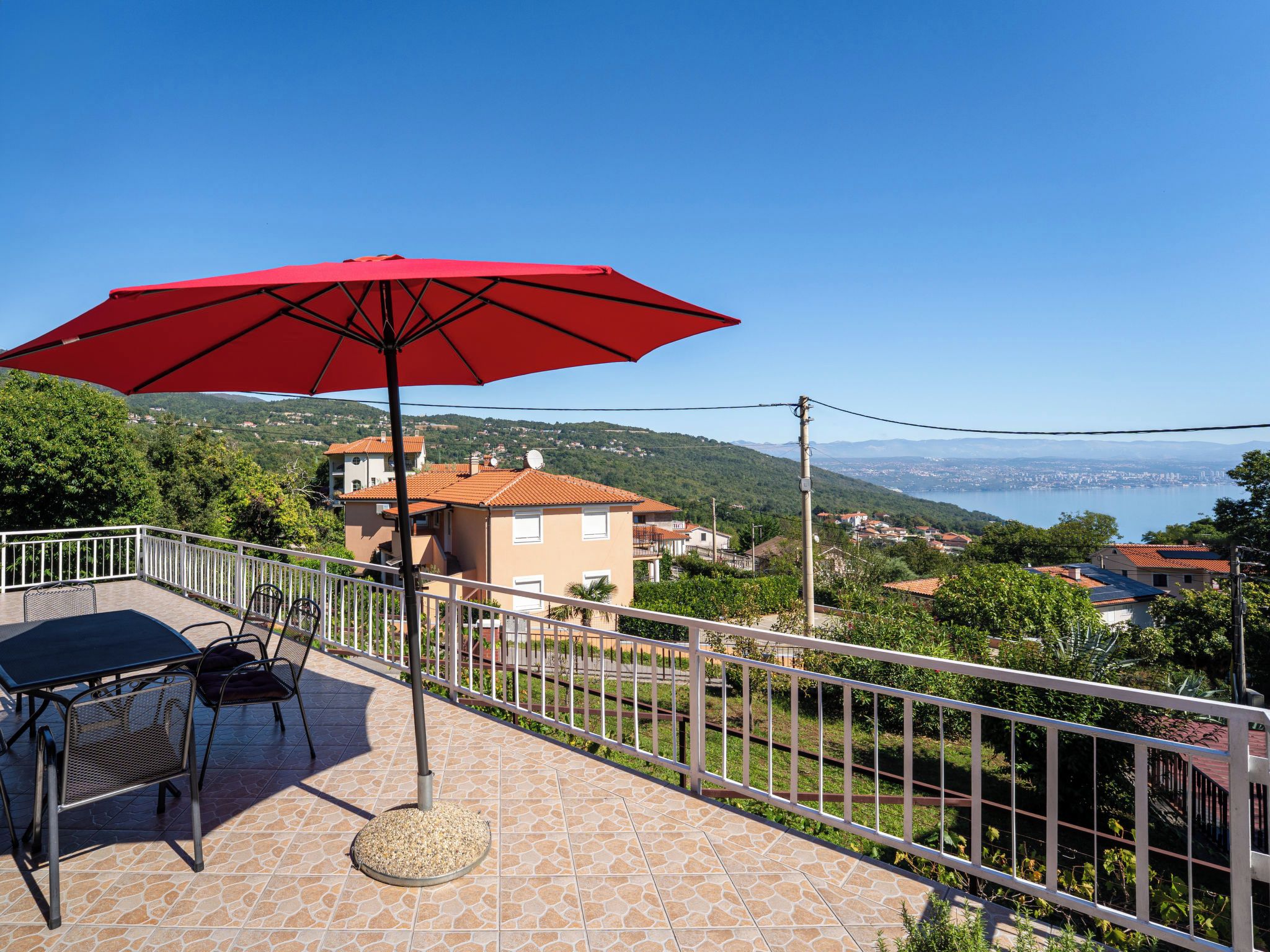 A terrace with a table and chairs, a red umbrella, and a view of a coastal town.