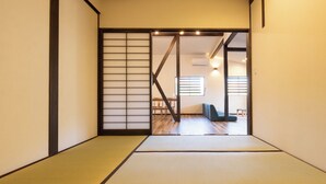 Une pièce de style japonais traditionnel, avec un sol recouvert de tatamis, des portes coulissantes et une vue sur un espace de vie comprenant un canapé et une table.