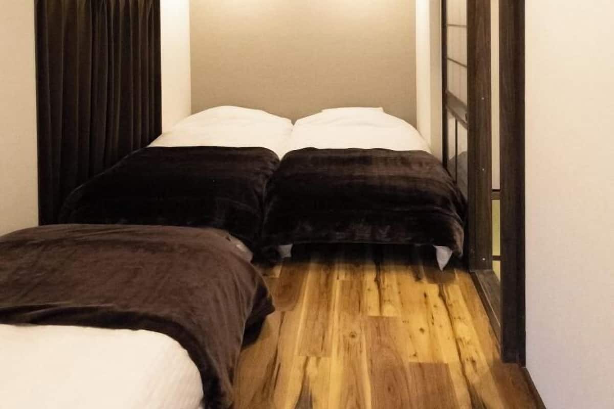 A narrow room with a wooden floor, two beds with dark bedding, and a window with a black frame.