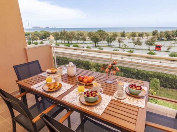A balcony table with breakfast items, a vase of flowers, and a view of the sea and parking area.