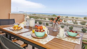 Una mesa en el balcón con alimentos para el desayuno, un jarrón de flores y vista al mar.