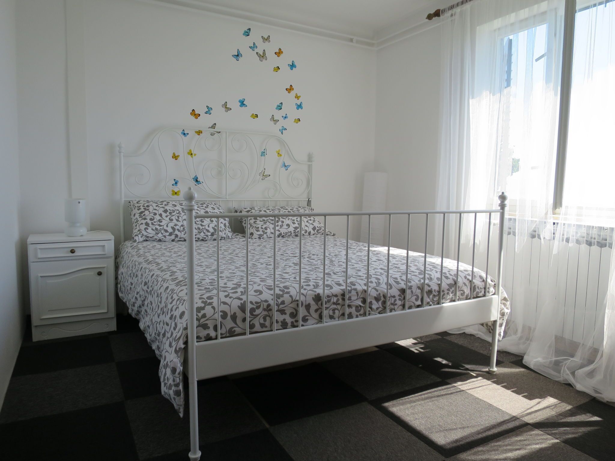 A white metal bed with a patterned bedspread, a bedside table with a lamp, and a window with sheer curtains.
