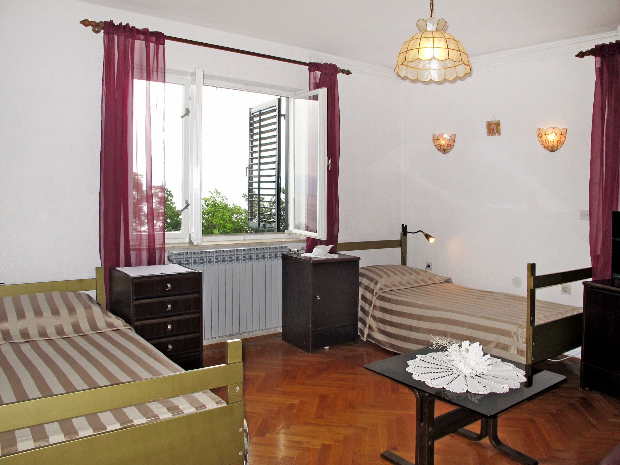 A bedroom with two beds, a dresser, a table with a lace tablecloth, a window with curtains, and wall sconces.