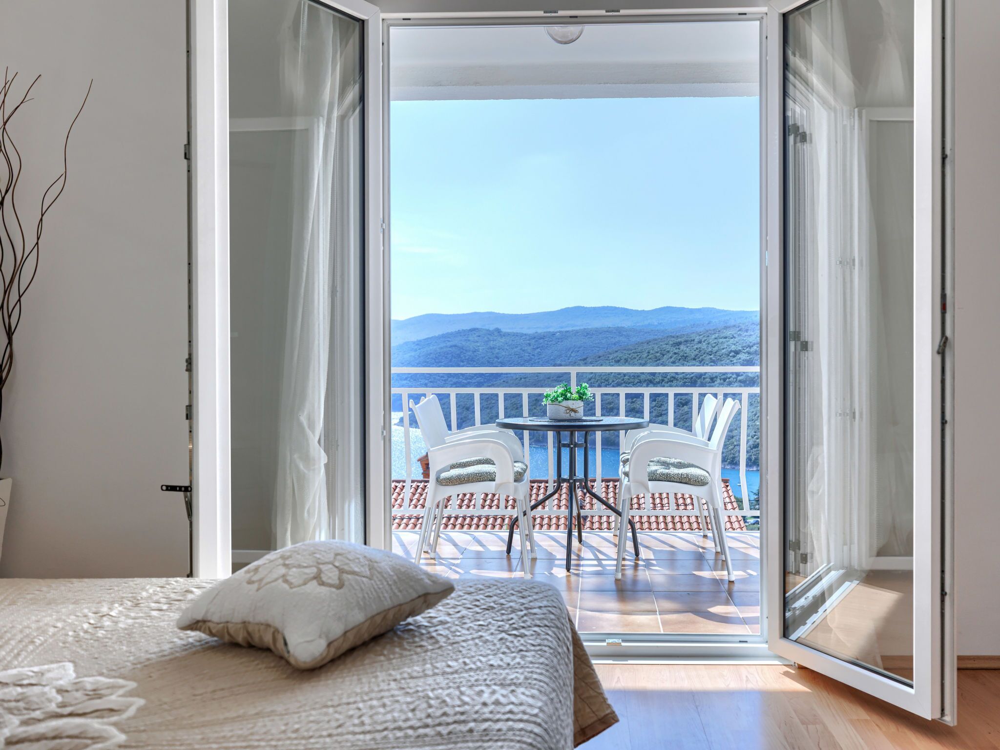 A bedroom with a large sliding glass door leading to a balcony with a table and chairs, overlooking a mountainous landscape.