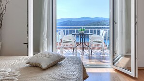 Quarto com uma grande porta de correr de vidro que dá para uma varanda com mesa e cadeiras, com vista para uma paisagem montanhosa.
