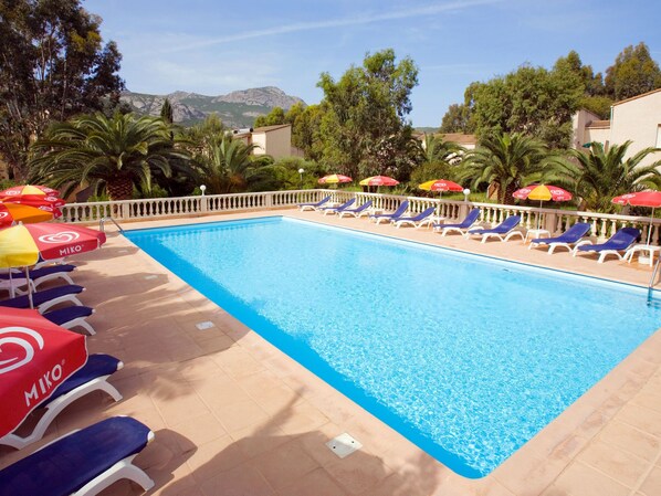 A swimming pool with sun loungers and umbrellas, surrounded by palm trees and mountains in the background.