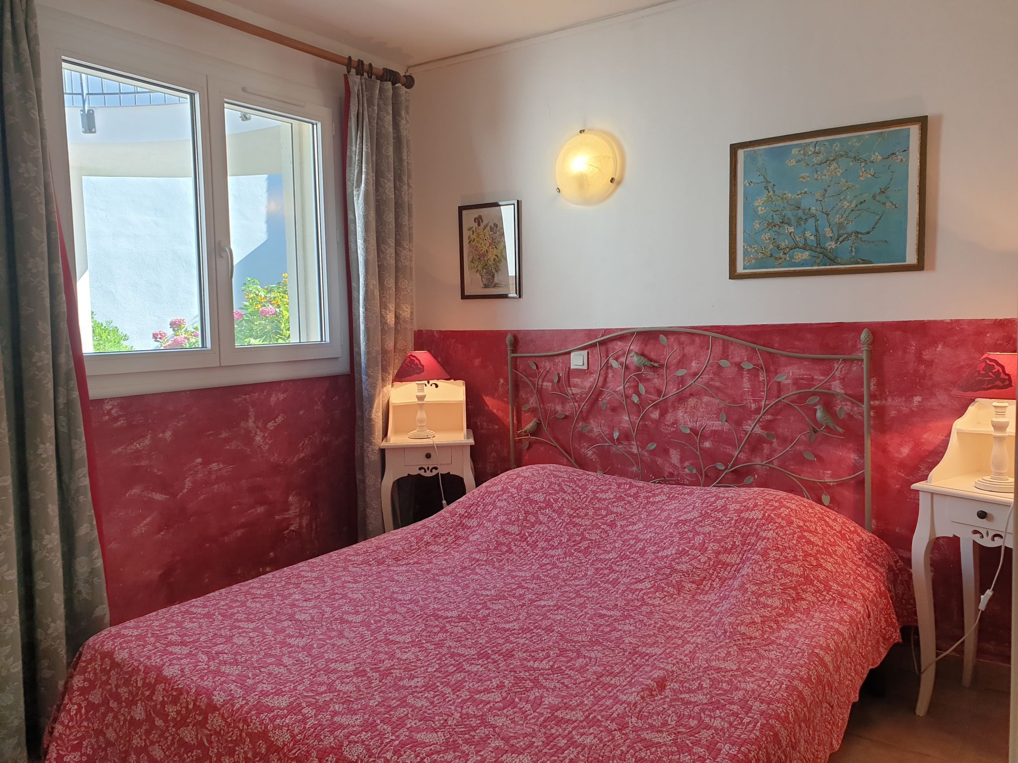 Une chambre à coucher avec un lit, une table de chevet, une lampe fixée au mur et une fenêtre avec des rideaux.